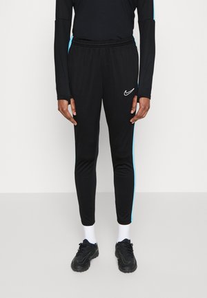 Person trägt schwarze Nike Trainingshose mit hellblauen Seitenstreifen, weiße Socken und schwarze Sportschuhe auf einfarbigem Hintergrund.