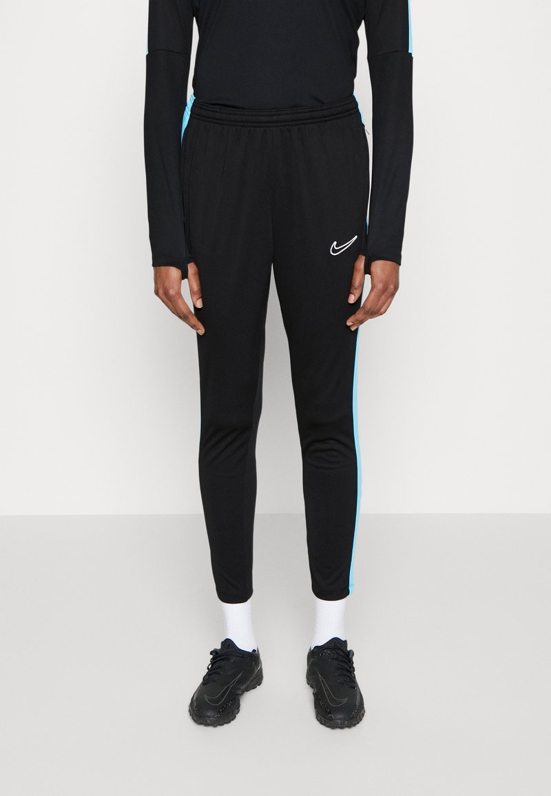 Personne portant un pantalon de survêtement noir Nike avec des bandes latérales bleu clair, des chaussettes blanches et des chaussures de sport noires sur un fond uni.