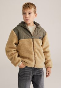 Barna testű polárdzseki zöld steppelt kapucnival. Elöl cipzáras, elasztikus ujjvégekkel, és feltűnő logófolttal.