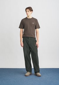 Brun bomulds t-shirt med "OBEY" logo, parret med mørkegrønne corduroy bukser og beige sko, stående på en blå tæppeoverflade.