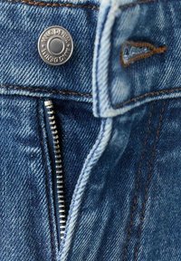 Pantalones vaqueros de mezclilla con cremallera plateada y botón metálico. La tela es azul descolorida con detalles de costura visibles.