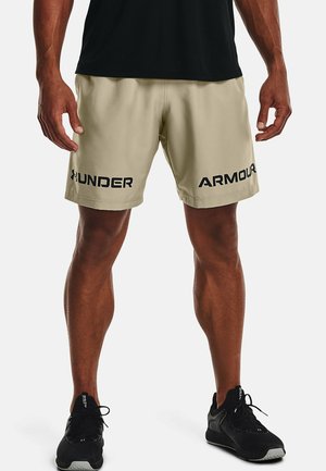 Mann trägt beige Under Armour-Shorts mit schwarzem Schriftzug auf den vorderen Oberschenkeln, schwarzes Shirt und schwarze Sportschuhe, steht vor weißem Hintergrund.