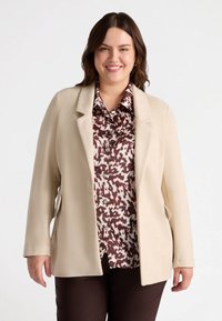 Blazer beige con un design strutturato, caratterizzato da due tasche frontali e revers a scollatura, indossato sopra una camicia a disegni marrone e crema.