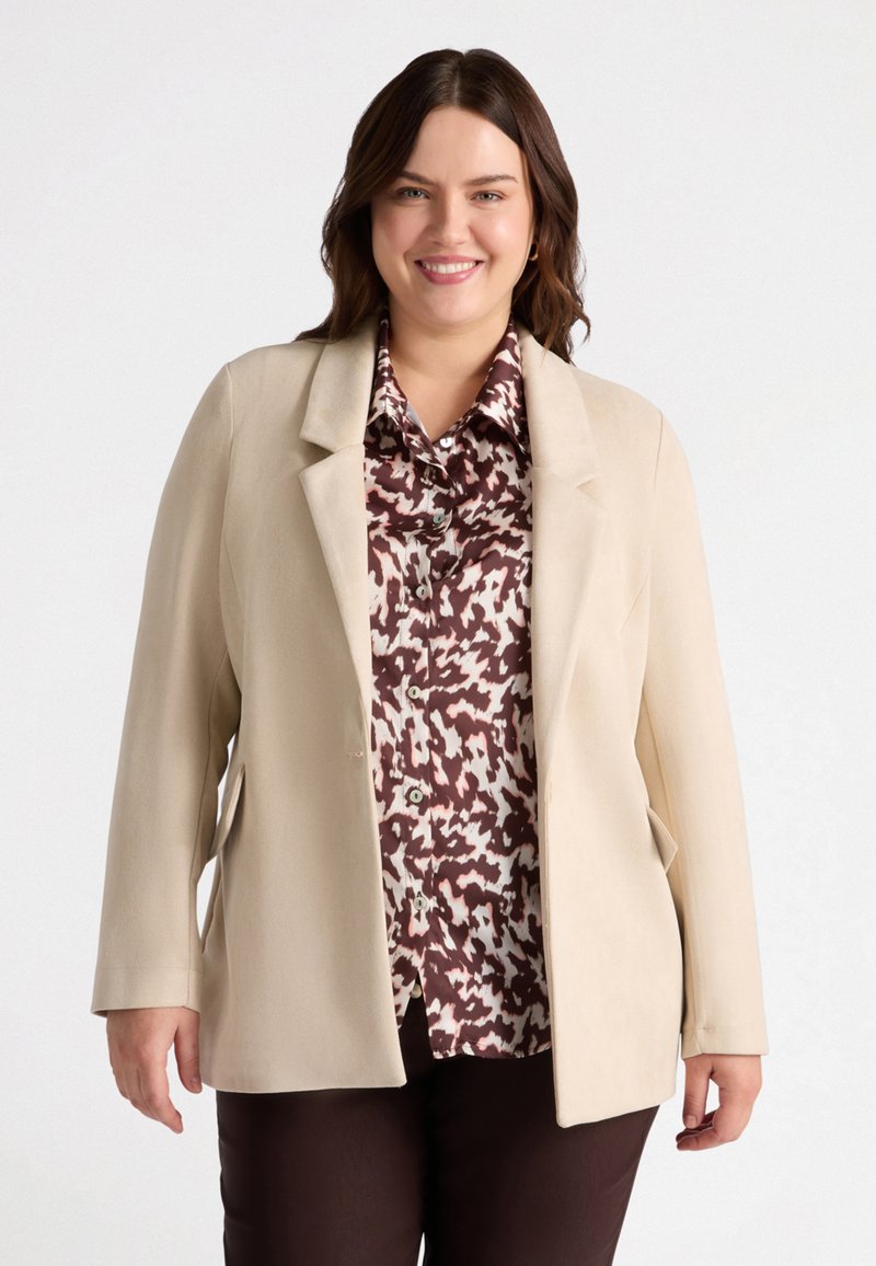 Blazer beige con un design strutturato, caratterizzato da due tasche frontali e revers a scollatura, indossato sopra una camicia a disegni marrone e crema.