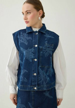 Jeune femme portant un gilet en jean bleu à motif floral sans manches avec des boutons argentés par-dessus un chemisier blanc à manches longues, regardant vers la gauche.
