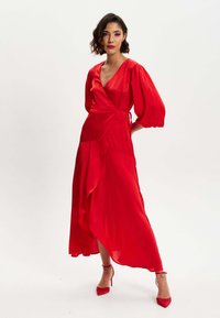 Red wrap dress with a deep V-neckline, puff sleeves, and an asymmetric hem. Made of smooth fabric, paired with red stiletto heels.