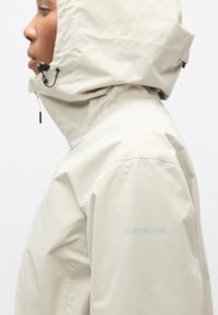 Veste imperméable beige avec capuche, dotée d'une surface texturée et de cordons de serrage ajustables. Logo "ICEPEAK" sur la manche gauche.
