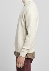 Maglione a collo alto color crema con una texture morbida, orlo e collare a coste. Indossato sopra una camicia marrone e abbinato a pantaloni bordeaux.
