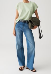 T-shirt in cotone verde chiaro con maniche arrotolate, jeans blu a gamba larga, cintura nera e borsa lavorata scura. Sandali con cinturini sottili.