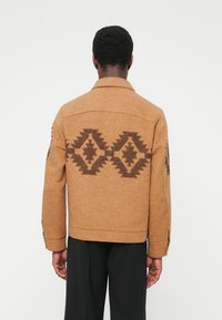 Brun uldjakke med et geometrisk mønster i mørk brun på ryggen, med krave og knappede manchetter.