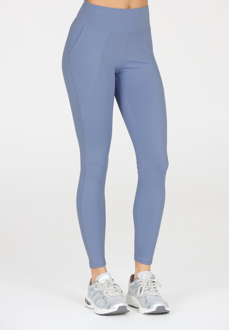 Höga, ljusblå leggings i stretchmaterial; har sidfickor och kontrasterande sömmar. Bärs med gråa sportskor.