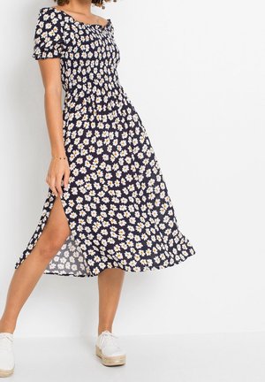 Donna che indossa un vestito midi blu navy con stampa di margherite bianche e gialle, maniche corte, spacco laterale e sneaker bianche con plateau.
