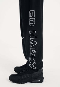 Svarta sweatpants har hög kontrastrik vit "ED HARDY" text längs benet. Kombinerade med svarta sneakers betonar outfiten en monochrom look.