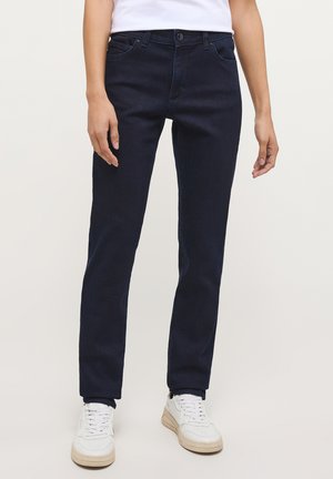Personne portant un jean bleu foncé ajusté et des baskets blanches, debout devant un fond clair uni.