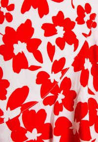 Rotes Blumenmuster auf weißem Stoff, gestaltet mit großen Blumen und Blättern. Auffällige Nähdetails entlang der Ränder.
