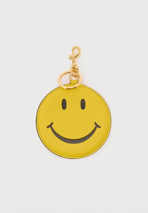 Llavero redondo amarillo con cara sonriente negra y clip metálico dorado junto a un anillo para llaves sobre un fondo blanco.