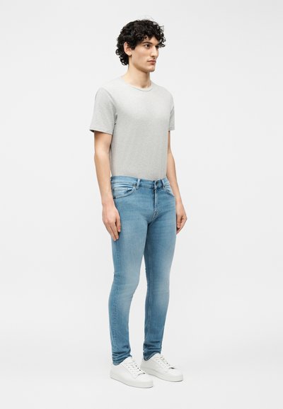 Νέος άνδρας με σγουρά μαλλιά, φορώντας ανοιχτό γκρι μπλουζάκι, ανοιχτό μπλε στενά τζιν και λευκά αθλητικά παπούτσια, στέκεται μπροστά σε ένα απλό φόντο.