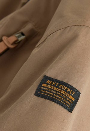 Beige fabric close-up featuring a black fabric label with yellow text: "NEXT SUPPLY," and details about authenticity and durability.
