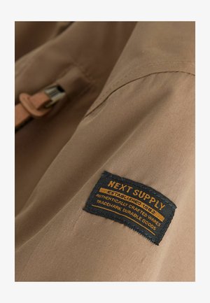 Smėlinis audinys artimas planas, kuriame matyti juoda audinio etiketė su geltonu tekstu: "NEXT SUPPLY" bei informacija apie autentiškumą ir patvarumą.