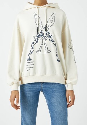 Persona con sudadera con capucha crema con un gráfico negro de Bugs Bunny llorando y vaqueros azules, manos relajadas a los lados sobre fondo blanco.