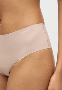 Γυναίκα με αδιάκοπα μπεζ εσώρουχα Calvin Klein, που δείχνει τη μέση και το πάνω μέρος του μηρού σε λευκό φόντο.