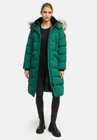 Zelena pufasta jakna s kapuco obloženo z umetnim krznom, z zadrgo na žepih in dolžino do kolen, v kombinaciji s črnimi hlačami in masivnimi črnimi škornji.