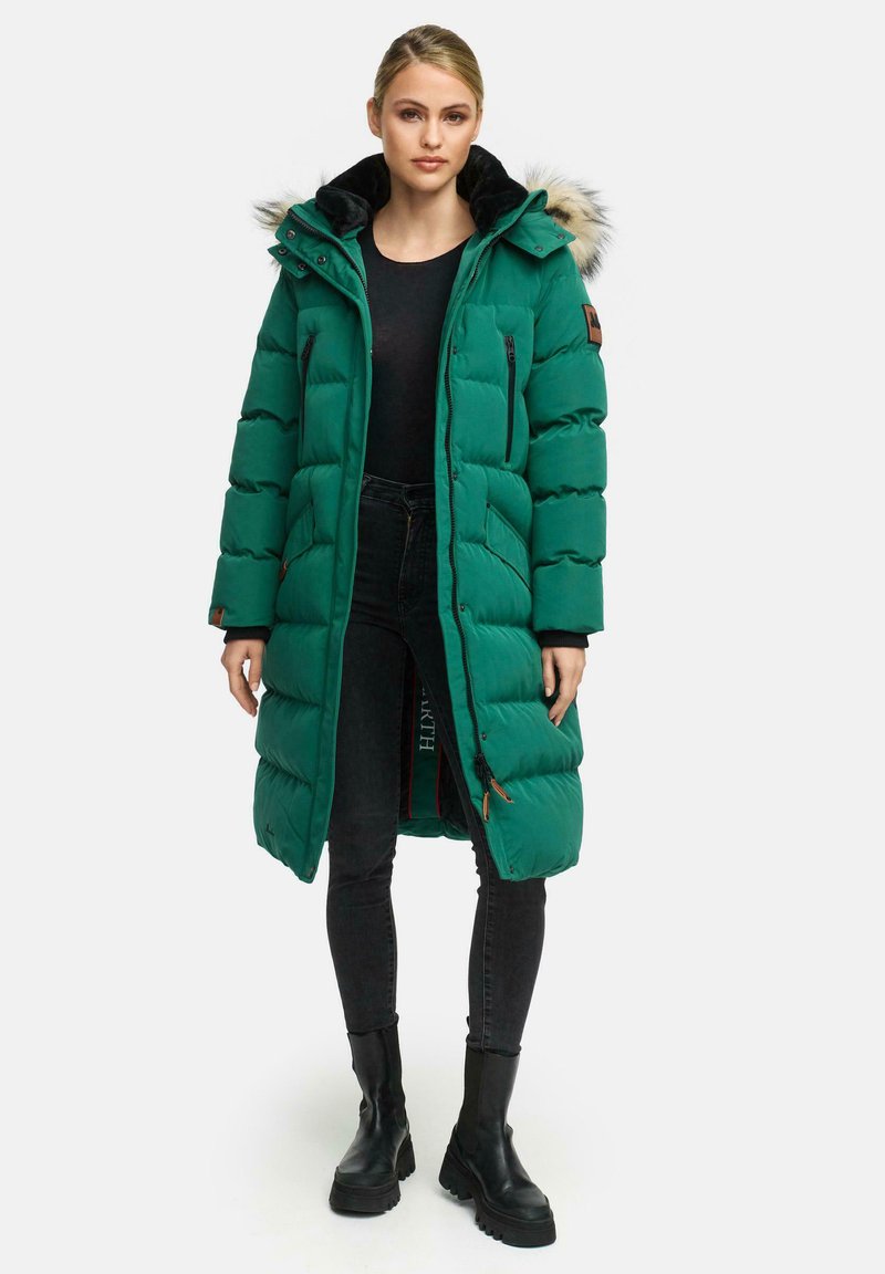 Zelena pufasta jakna s kapuco obloženo z umetnim krznom, z zadrgo na žepih in dolžino do kolen, v kombinaciji s črnimi hlačami in masivnimi črnimi škornji.