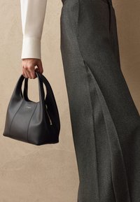 Schwarze Ledertasche mit zwei Griffen und einem Reißverschlussdetail. Sie hat eine glatte Textur und eine runde Form. Kombiniert mit maßgeschneiderten grauen Hosen.