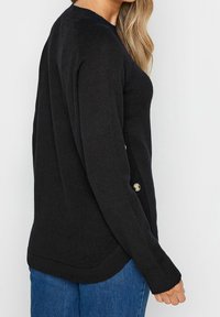 Svart stickad tröja med långa ärmar, med en sidoknappdetalj och rundad nederkant. Bärs med blå jeans.