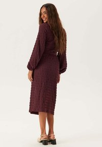 Bordeaux langarmige jurk van gestructureerde stof, met een ceintuur in de taille en midi-lengte, gecombineerd met sandalen met blokhak.