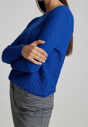 Blauwe gebreide trui met een diamantpatroon, voorzien van een ronde halslijn en ribgebreide boorden, gecombineerd met een geblokte broek.