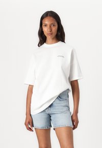 White cotton t-shirt with short sleeves, round neckline, and subtle black text on the front. Paired with light blue denim shorts.