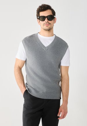Gilet grigio lavorato a maglia con scollatura a V, caratterizzato da una texture a costine e maniche corte, indossato sopra una maglietta bianca, abbinato a pantaloni scuri e occhiali da sole.