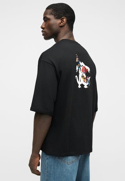 Άνδρας που φοράει μαύρο φαρδύ μπλουζάκι με λευκό και πολύχρωμο λουλουδένιο σχέδιο στην πλάτη, συνδυασμένο με ανοιχτό μπλε τζιν.