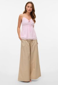 Šviesiai rožinė kamisolė su nėrinių apdaila ir reguliuojamomis petnešėlėmis, derinama su smėlio spalvos plataus pjūvio kelnėmis su aukštu liemeniu ir klostėmis.