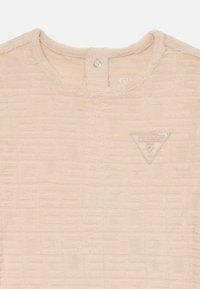 Beige teksturert kortermet topp med et geometrisk mønster, rund hals og en trekantet logo-patch på brystet.