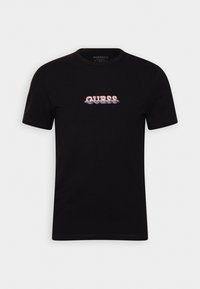 Camiseta negra de manga corta hecha de algodón. Presenta un logo central de "GUESS" en rojo y azul, con una textura suave y un cuello redondo.