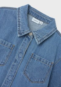 Veste en denim bleu clair avec un col classique, deux poches poitrine, des coutures cuivrées et une étiquette de marque à l'intérieur du col.