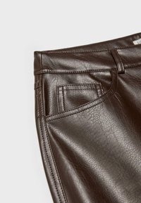 Gros plan sur un pantalon marron foncé en similicuir montrant les coutures, la ceinture, les passants de ceinture, la poche avant et la surface texturée.