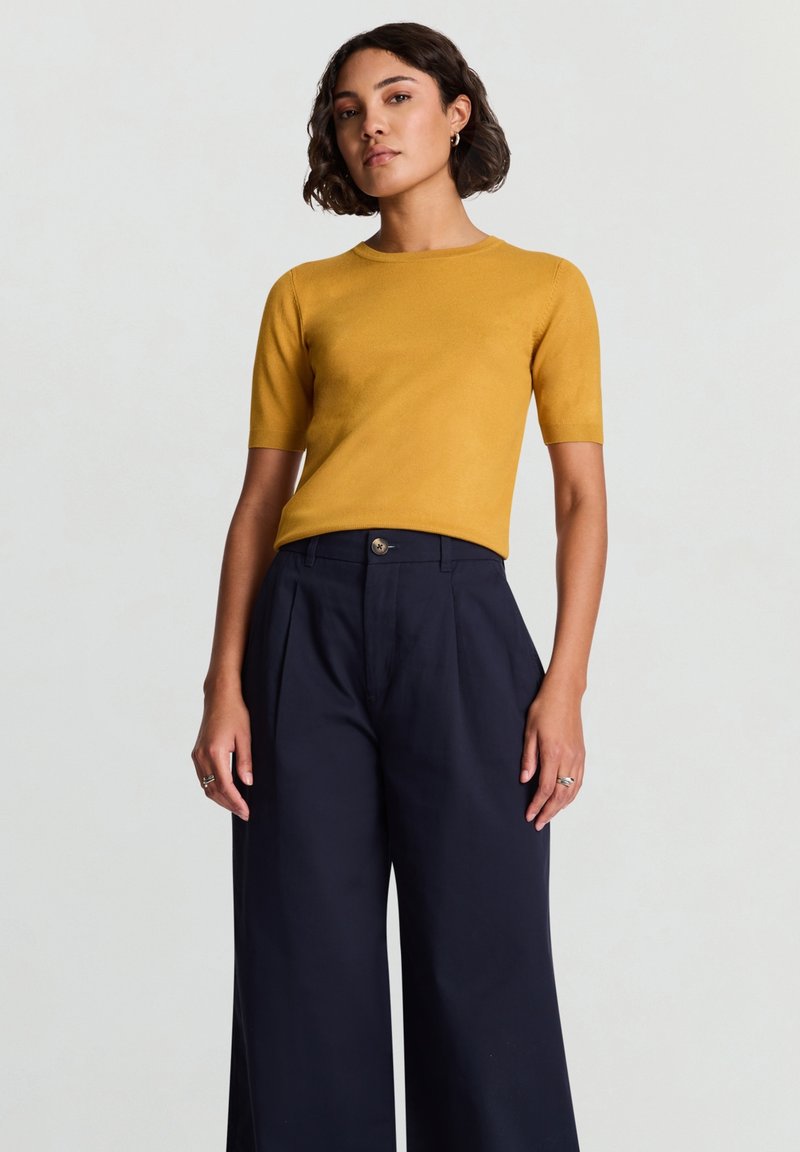 Mustard short-sleeve top paired with high-waisted, wide-leg navy trousers, featuring a smooth texture and button detail at the waist.