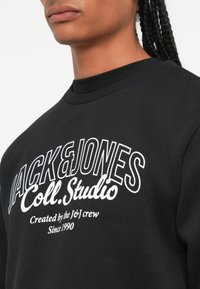 Čierna mikina so žebrovaným výstrihom a bielou potlačou textu s nápisom "JACK&JONES Coll. Studio" a "Created by the J&J crew Since 1990."