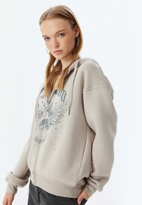 Sudadera con capucha y cremallera en beige claro, con un estampado gráfico que incluye alas, estrellas y texto; fabricada en un tejido suave y texturizado.