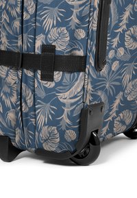 Maleta de tela azul con un patrón de hojas tropicales beige, con cremalleras negras y cuatro ruedas negras para facilitar la movilidad.