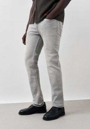 Uomo che indossa jeans slim fit grigio chiaro, mocassini neri e una camicia scura con le mani nelle tasche, in piedi su un pavimento grigio semplice.
