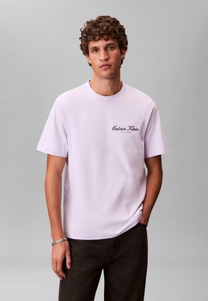 Man with curly hair wearing a light purple Calvin Klein T-shirt and black pants, standing with one hand in his pocket against a plain gray background.