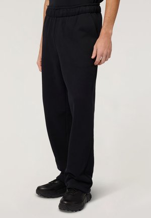 Persoon die zwarte joggingbroek met elastische taille en zwarte sneakers draagt, staand tegen een neutrale achtergrond, onderlichaam zichtbaar.