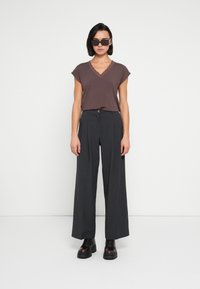 Top marrón de cuello en V con ribete de encaje, pantalones grises de pierna ancha y botas negras gruesas; la modelo lleva gafas de sol negras extragrandes.