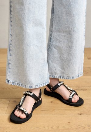 Black sandals feature a T-strap design with faux pearl and crystal embellishments, on a flat rubber sole, paired with light denim wide-leg pants.