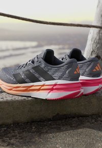 Chaussures de course grises et noires avec des semelles coussinées orange et rose posées sur un rebord à l'extérieur près d'une corde et d'un arbre.