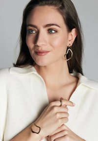 Biżuteria w kolorze złota z organicznymi kształtami: duże kolczyki, bransoletka o otwartym wzorze i teksturowany pierścionek na tle neutralnego swetra.